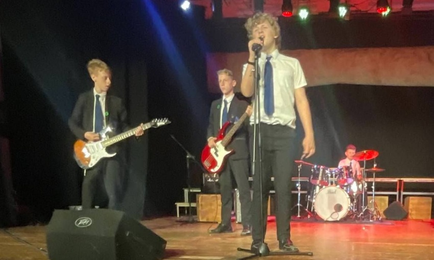 Image shows KS3 pupils performing music on stage - there are 4 pupils, 1 on drums in the background, 2 on electric guitar and 1 singing at the front.